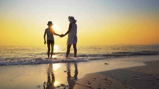 一家人在海滩上散步，去海边日落，溅起海浪。 女人穿白色连衣裙，草帽男孩戴帽子