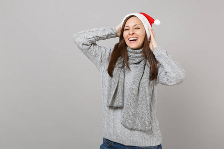 微笑的圣诞老人女孩，戴着灰色毛衣围巾，戴着圣诞帽，双手放在头上，孤立在灰色背景的工作室肖像上。 新年快乐，2019年庆祝节日聚会