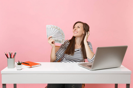 年轻的梦想微笑的女人，想着抬起头，拿着一捆很多美元的现金，在白色的桌子上工作，电脑笔记本电脑孤立在粉彩背景上。 成就事业。 复制
