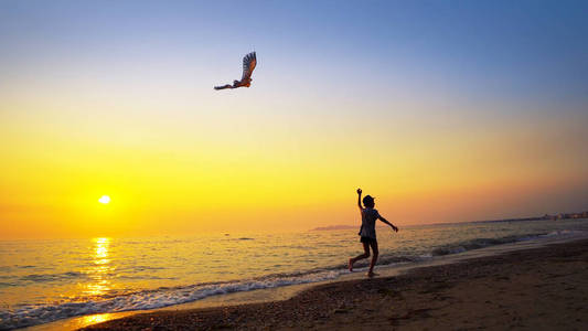 活力带带着帽子的小男孩站在海边,在充满活力的夕阳下玩着放风筝照片