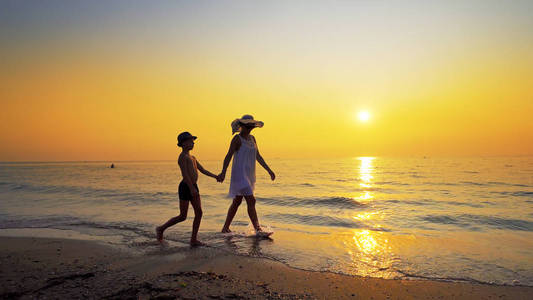 幸福的家庭走在海边，在日落时，漫延的水踢着海浪。 母亲穿白色连衣裙，两人都戴帽子
