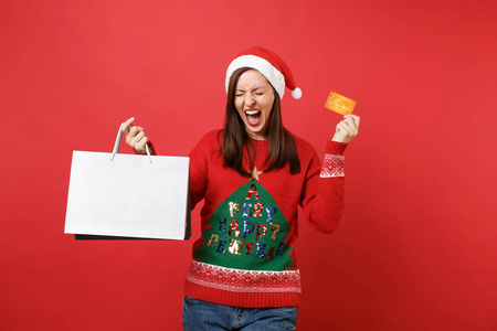 欣喜若狂的年轻圣诞老人女孩尖叫，闭着眼睛，拿着信用卡包袋，购物后，孤立在红色背景下。 新年快乐2019庆祝节日派对概念