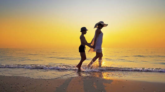 幸福的家庭走在海边，在日落时，漫延的水踢着海浪。 母亲穿白色连衣裙，两人都戴帽子