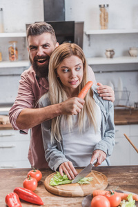 有趣的男人把胡萝卜放在女人的喉咙里，而女孩在做饭