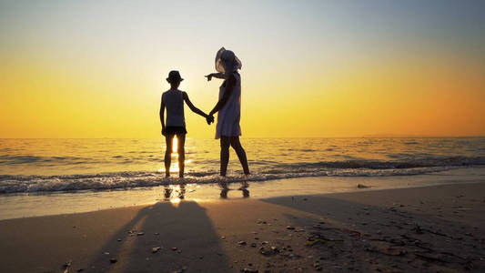一家人在海滩上散步，去海边日落，溅起海浪。 女人穿白色连衣裙，草帽男孩戴帽子
