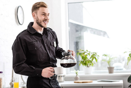 微笑的男人穿着警服从厨房窗户附近的水壶里倒咖啡
