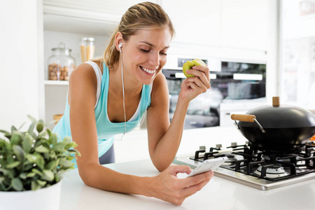 一位美丽的年轻运动女子在厨房吃苹果时,一边听音乐,一边用智能手机发