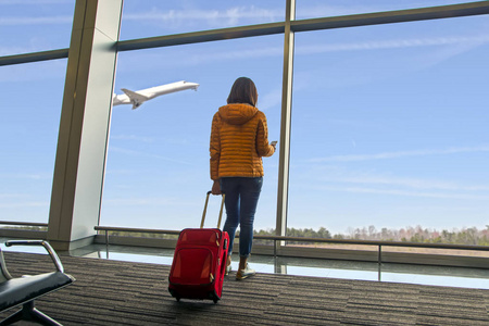 一位年轻漂亮的旅行者女士在机场窗口看到飞机，女孩游客拿着智能手机，拿着红色的包，在大厅的飞机起飞前等待。 旅游度假及旅游概念