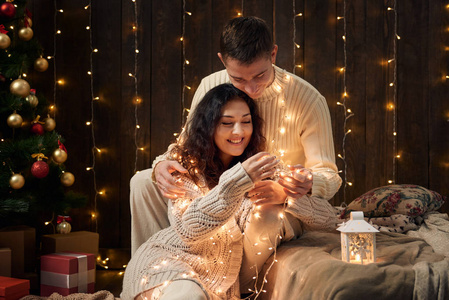 这对年轻夫妇穿着白色杉树的圣诞灯和装饰，穿着深色木制背景的寒假概念
