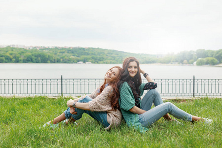 微笑的黑发和红色的女孩背靠背坐在草地上,看着镜头腿微微弯曲的膝盖.