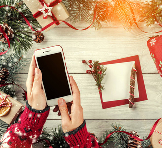 女性手拿手机。 圣诞礼品盒与红色装饰杉树枝松果纸卡备注假日背景。 圣诞节和新年快乐卡。 平躺式顶部视图