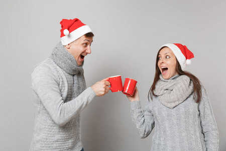 有趣的情侣女孩穿着红色圣诞帽，灰色毛衣，围巾，拿着一杯茶，孤立在灰色墙壁背景工作室的肖像上。 新年快乐，2019年假日派对理念。
