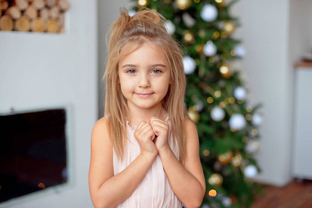 快乐的小女孩穿着豪华的连衣裙，双手靠近脸，背景上有五颜六色的灯光和圣诞树。 圣诞节，新年