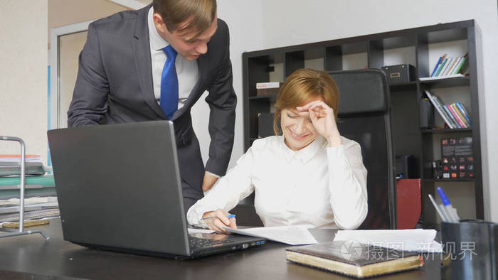 两个上班族坐在办公桌前, 一个女人在电脑上工作, 一个男人就在附近.