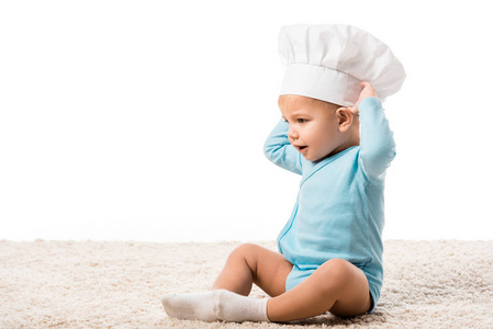 戴着厨师帽子的蹒跚学步的男孩，双手抱着头坐在地毯上，与白色隔离