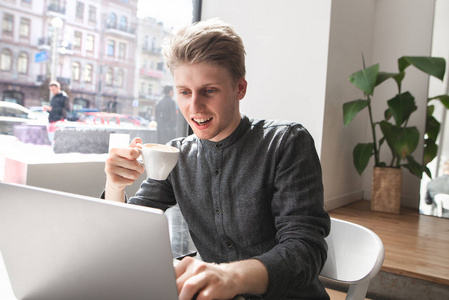 快乐的年轻学生坐在一家清淡凌乱的咖啡馆里，手里拿着一杯咖啡，用笔记本电脑，微笑着。 开朗的年轻人在咖啡馆里用笔记本电脑喝咖啡