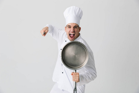 情感的年轻人厨师在室内隔离在白色的墙壁背景上,拿着煎锅