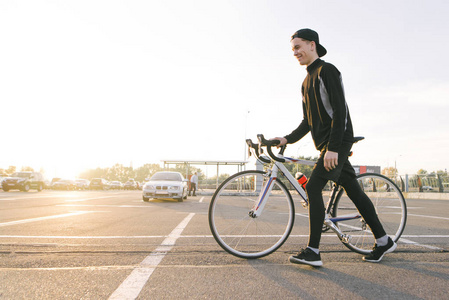快乐的年轻人穿着深色运动服和帽子，在夕阳的背景下在街上拉着一辆自行车，微笑着。 日落时骑自行车的人。