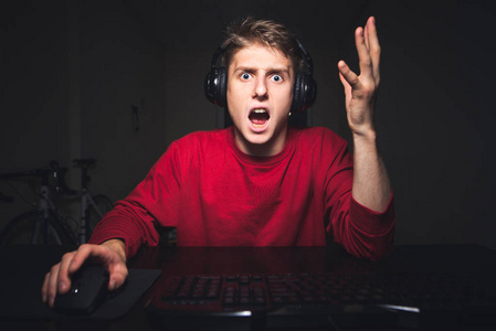 年轻的玩家对电子游戏中的坏球队感到愤怒。 一个情绪化的玩家在家里玩网络电子游戏的肖像，在电脑上举起双手，从惊喜中举起。