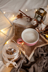 木制托盘上的热卡布奇诺杯在床上。 舒适的装饰。 早餐。 杯子格子棉蜡烛。 书。 圣诞灯。 假日。