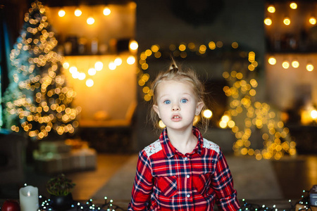 一个漂亮的小眼睛金发女孩坐在一个有圣诞树的房子里的木桌上，等待她的圣诞礼物。 圣诞节早晨概念
