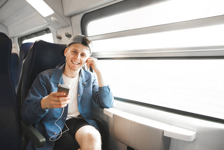 快乐的人上火车听音乐耳机里拿着手机看着相机微笑。微笑的年轻乘客坐在靠近窗户的火车上听音乐
