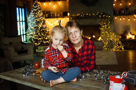 一位年轻的祖母和她的小孙女坐在桌子上吃圣诞礼物中的糖果。 圣诞节家庭早晨的概念。