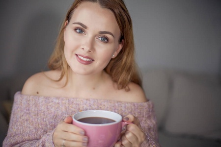 年轻漂亮的女人拿着一大杯粉红色的茶。 穿针织毛衣的女孩喝咖啡。 很舒服。 秋天。 春天。 冬天。