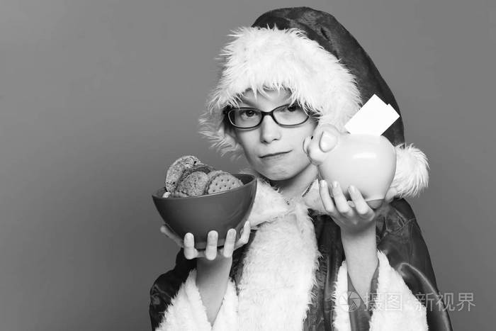 年轻可爱的圣诞老人男孩与眼镜在红色毛衣和新年圣诞帽子举行粉红色小猪猪银行和巧克力臀部饼干在碗中的蓝色工作室背景