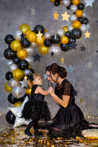 女权力党与美丽的模特母亲和可爱的宝贝女儿穿着通风的黑色花式礼服庆祝爱与金色的星星和空气栏杆。由摄影师设计的枕头星