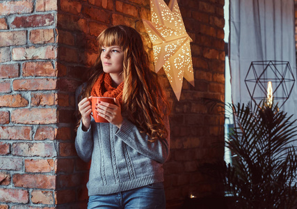 一个孤独的红头发的女孩在靠在砖墙上拿着一杯咖啡的温暖毛衣的肖像