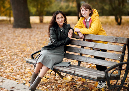 两个女孩在秋天的城市公园。他们坐在长凳上聊天。