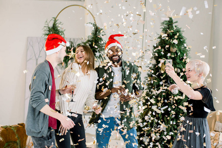 穿着时髦服装的年轻人站在圣诞树前拿着一杯香槟，一边笑着看着掉落的金色纸屑