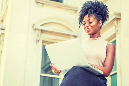 通往成功的道路。 年轻的非洲裔美国妇女，发型是非洲式的，穿着无袖浅色上衣，坐在纽约的老式办公楼旁，低头在笔记本电脑上工作，微笑着