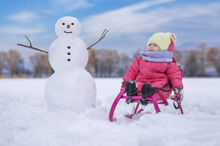 可爱的孩子女孩在明亮的雪地上，在雪人附近的粉红色雪橇上。 冬季户外活动