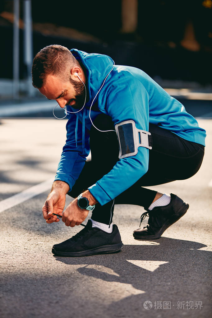 男性跑步者蹲着系鞋带耳朵耳机上的运动服健康的生活方式概念