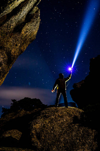 在岩石上的人用一个非常有力的手电筒照亮黑暗的夜空照片
