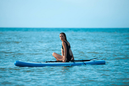 愉快美丽的年轻女孩与划桨板在海滩上。背景中的蓝色大海。暑假理念