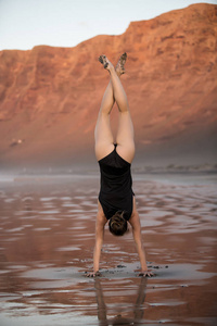 适合女性的后视图，展示倒立，同时在海滩上做瑜伽，站在潮湿的沙子上