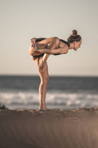 站在沙滩上的一条腿上做瑜伽体式的开朗肌肉女人的侧视图