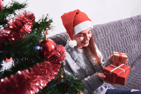 年轻快乐的女人，穿着毛衣，戴着红色圣诞帽，坐在沙发上，拿着圣诞礼物。新年和圣诞节假期的概念。