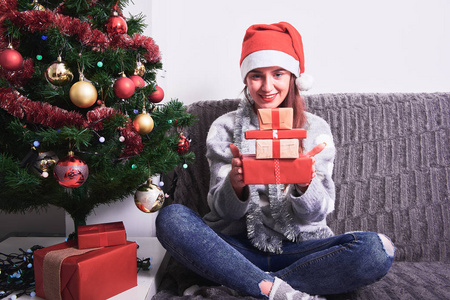年轻快乐的女人，穿着毛衣，戴着红色圣诞帽，坐在沙发上，拿着圣诞礼物。新年和圣诞节假期的概念。