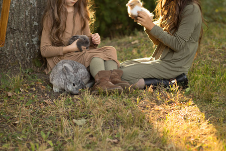 两个女孩坐在草地上的一棵树下，抚摸着小兔子，在附近的一个盒子里抱着一只灰色的大兔子。 米色和绿色的柔和色调。秋天或春天