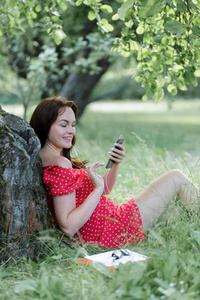 年轻漂亮的女人坐在树下的草地上，用她的智能手机