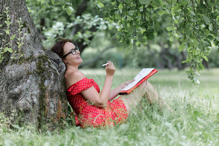 年轻女子坐在树下的草地上写笔记