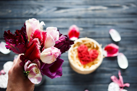 女人的手拿着一束鲜花郁金香和红色的木制心躺在黑暗的桌子上，周围有粉红色紫色的花瓣。情人节母亲节浪漫概念