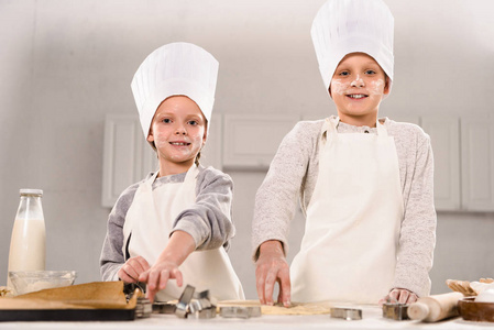 快乐的孩子们戴着厨师帽和围裙，在厨房的桌子上切面团做饼干