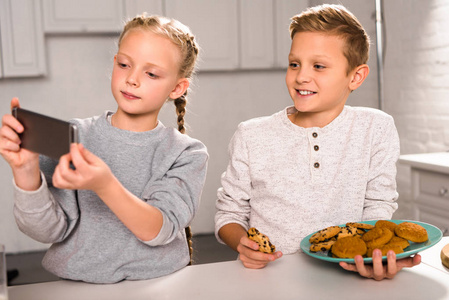 可爱的孩子在智能手机上自拍，弟弟拿着盘子，厨房里有美味的饼干。