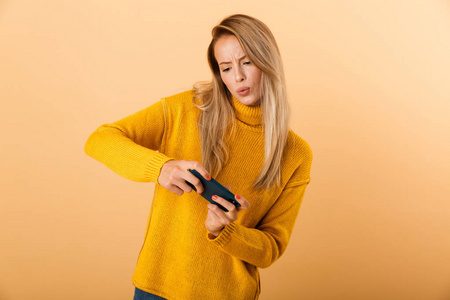 一位穿着毛衣,站在黄色背景下玩手机游戏的快乐年轻女子的肖像.照片