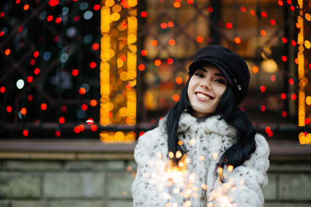 令人惊叹的年轻女子在模糊的城市灯光下用闪闪发光的光庆祝新年。 空的空间